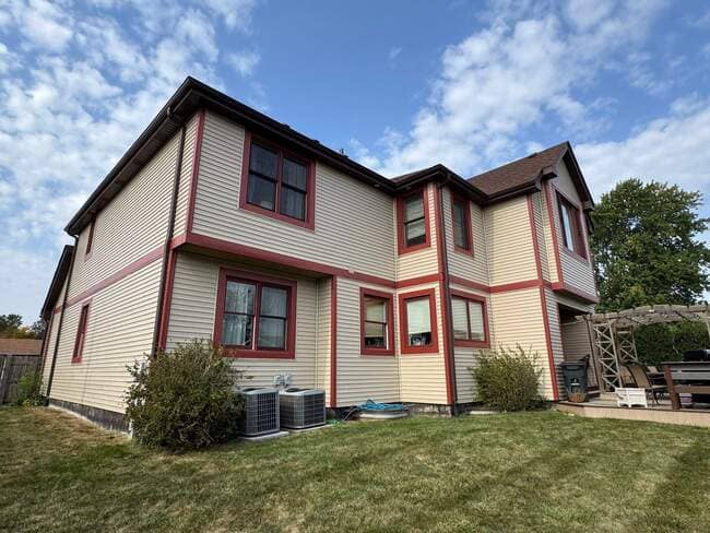 Ground-level view of a two-story residential home featuring newly installed tan vinyl siding with striking red/maroon trim around the windows and corners. The image highlights the clean lines of the new siding and the contrasting brown roof, showing a comprehensive exterior renovation under a blue and cloudy sky.